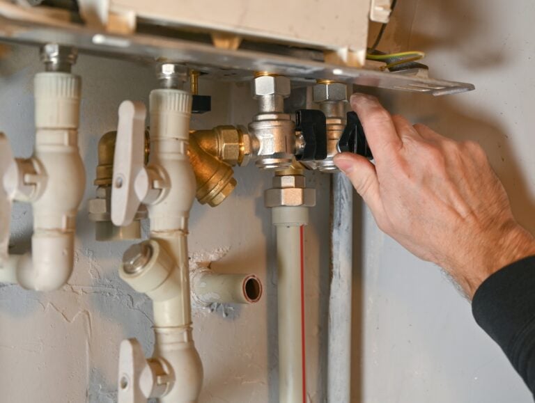 A human hand operates a black-handled valve within a complex plumbing manifold, featuring white plastic pipes, brass, and chrome fittings