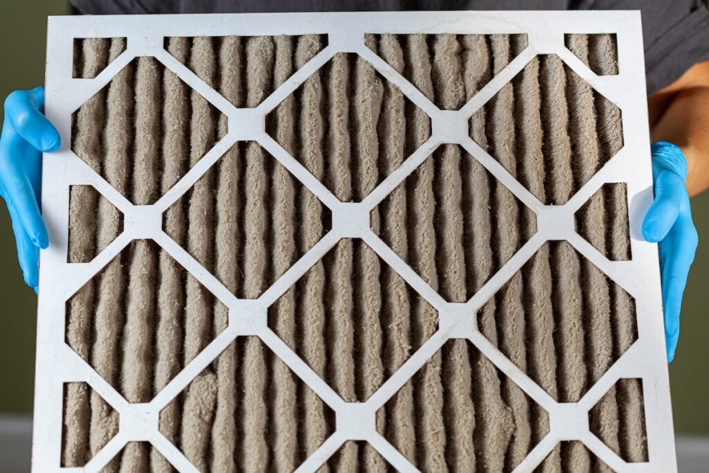 A person wearing blue gloves is holding a heavily clogged dirty air filter in hands before replacing it with the new one.