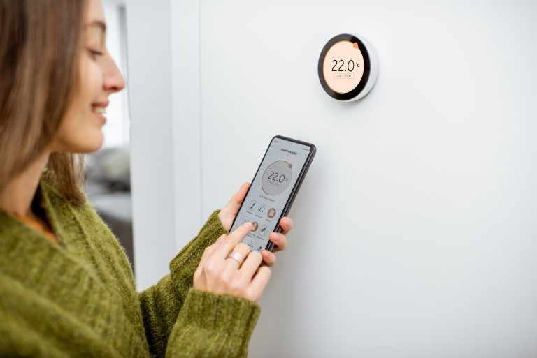 programmable thermostat installation Woman dressed in green sweater regulating heating temperature with a modern wireless thermostat and smart phone at home. Synchronization of thermostat with mobile devices concept
